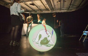 Photography of an exhibition space. On the floor there is a round disc on which the work ""Hassenberg"" is depicted. A person can be seen lying with his belly on a kind of ball and coloured smoke rises from the person's ears.