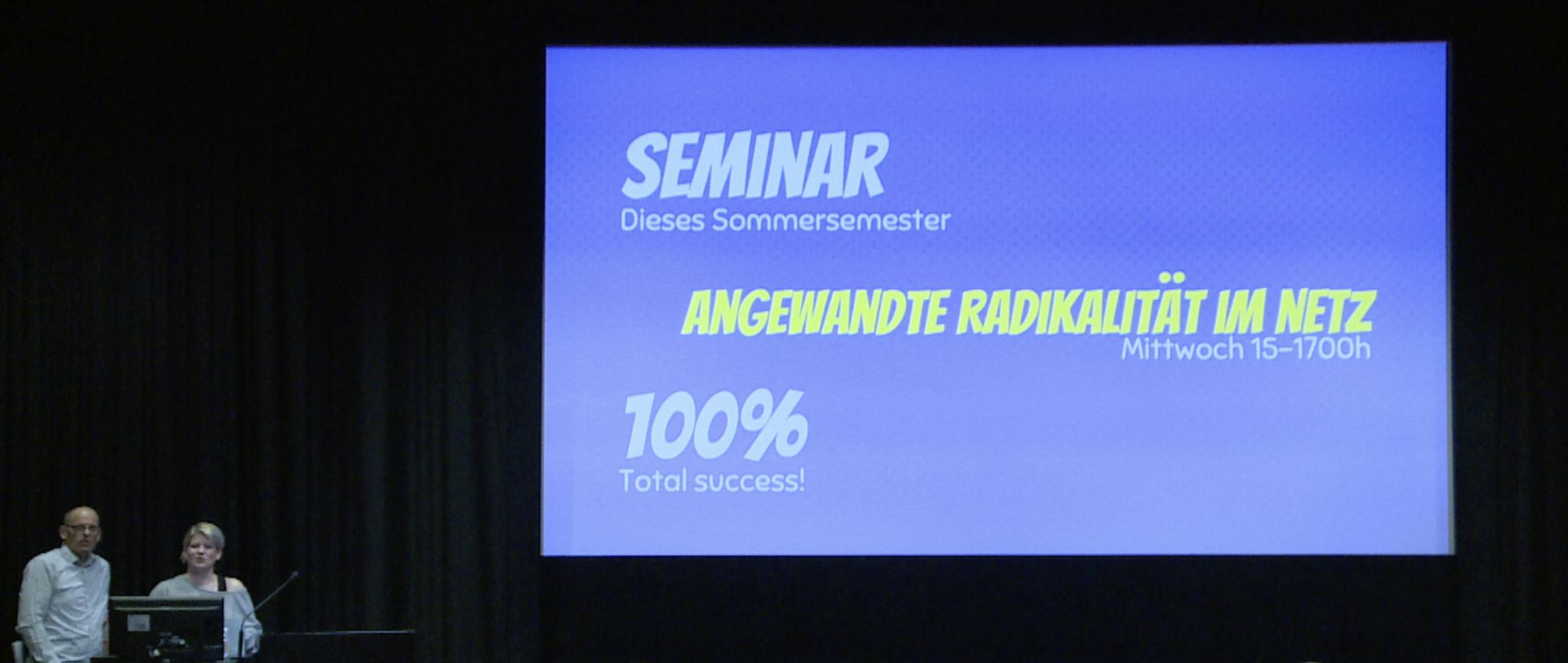 Photography of the inaugural lecture by the artist duo Utbmorgen, in the lower left corner you can see Prof. Hans Bernhard and Prof. Liz Haas. Also a projection in the middle of the picture with the title of the event: Angewandte Radikalität im Netz (Applied Radicality on the Net).